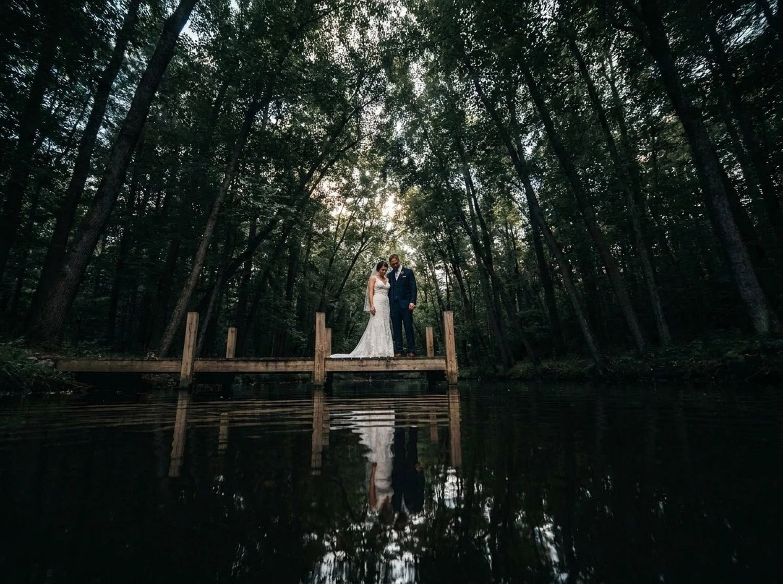 Bride and groom on a jetty surrounded by tall trees reflected in still water