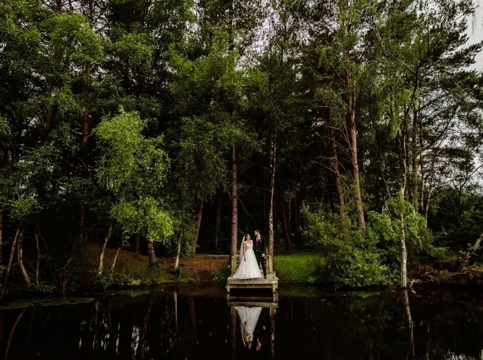 Bride and groom on a wooden jetty beside a woodland pond